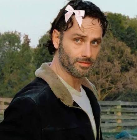 A bearded man with dark hair wears a pink bow in his hair and a black jacket with a fur collar, standing outdoors.