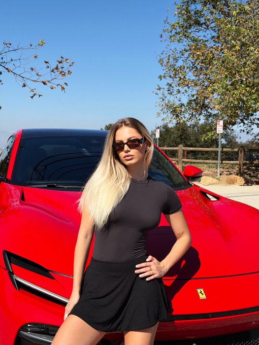 A blonde woman wearing sunglasses and a black outfit poses in front of a red Ferrari, with a wooden fence and trees visible in the background.