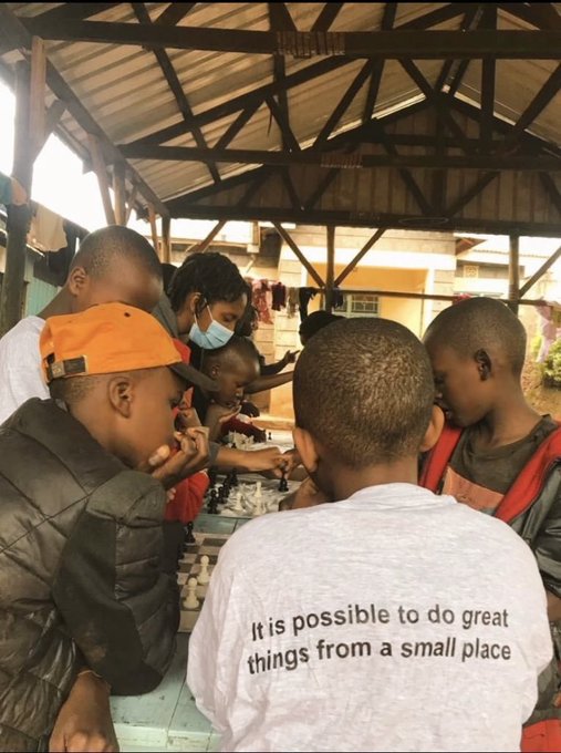 A group of people, including children and a person in a face mask, gathered around a table playing chess under a shelter; one person wears a white shirt reading 'It is possible to do great things from a small place.'