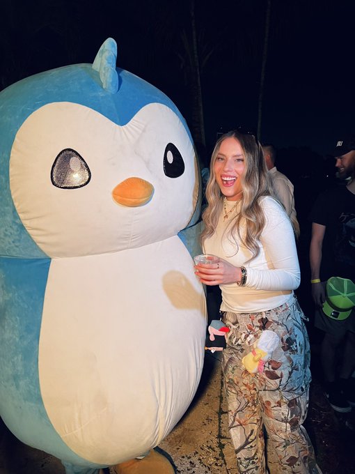 A smiling woman poses next to a large, plush Penguin-style mascot character at what appears to be a nighttime event, playfully connecting to the $PENGU token reference in the post. Her trendy camouflage pants and the colorful glow stick accessory complement the fun, festive vibe of the crypto community gathering.