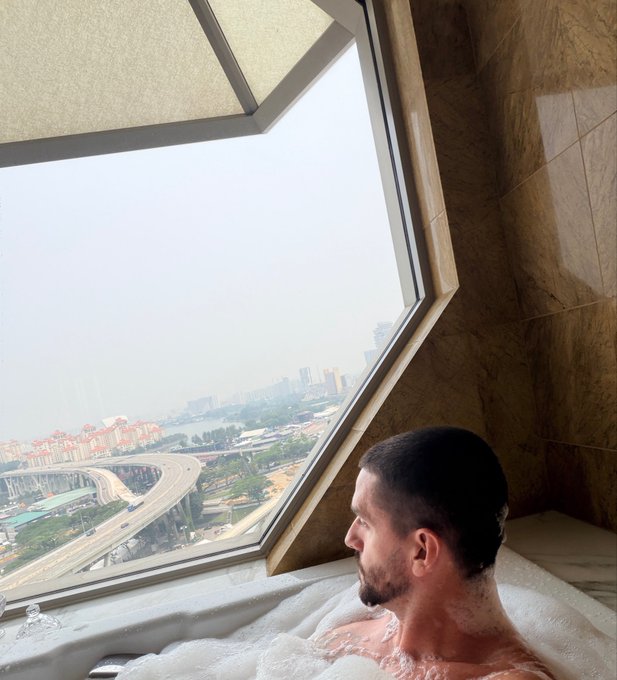 A man with a freshly shaved head relaxes in a bubble bath, gazing out a large angular window at a hazy cityscape with highways and high-rises in the distance. The luxurious bath view contrasts with the post's bullish Bitcoin price prediction, humorously pairing the "to the moon" mindset with a literal high-rise perspective.