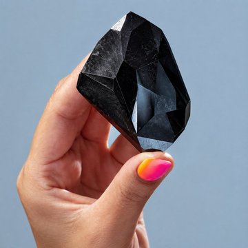 A hand with vibrant pink and yellow gradient nail polish holds up a large, faceted black diamond against a clear blue sky, showcasing the Black Enigma diamond owned by PulseChain and $HEX founder Richard Heart.
