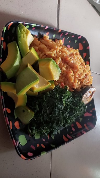 The image shows a plate of food consisting of rice, avocado slices, spinach leaves, and other vegetables arranged neatly on a dining table. The plate appears to be black with colorful designs around its edges, adding visual appeal to the presentation. The arrangement of the food items suggests that they are meant to be enjoyed together as part of a meal.