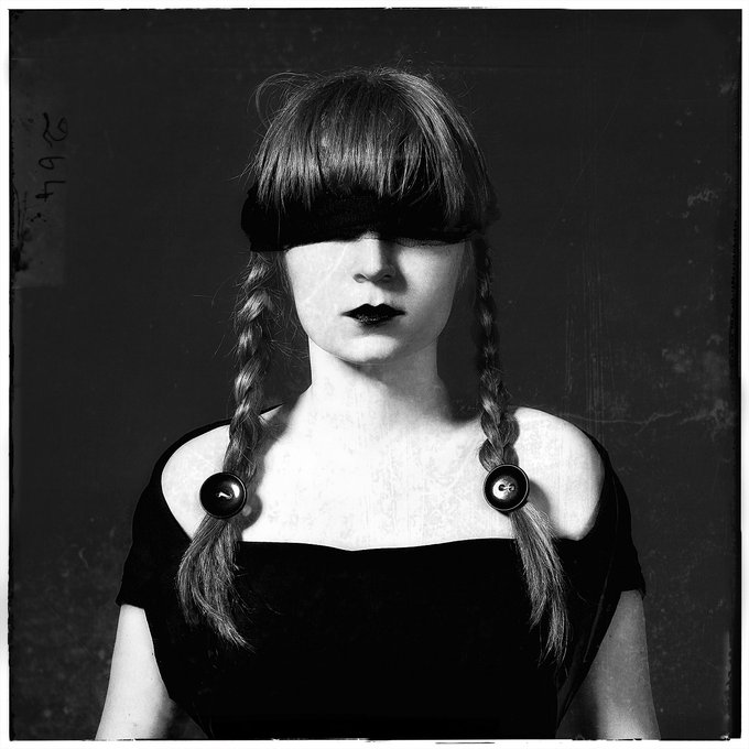 A black and white portrait of a woman with dark braided pigtails and a black blindfold covering her eyes, wearing a black top with button details, with the number '2967' visible on the left side.