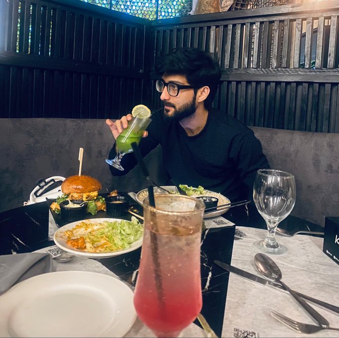 A young man with glasses and a beard sits at a restaurant table drinking a green beverage, with a burger, salad, and a pink cocktail visible on the table.
