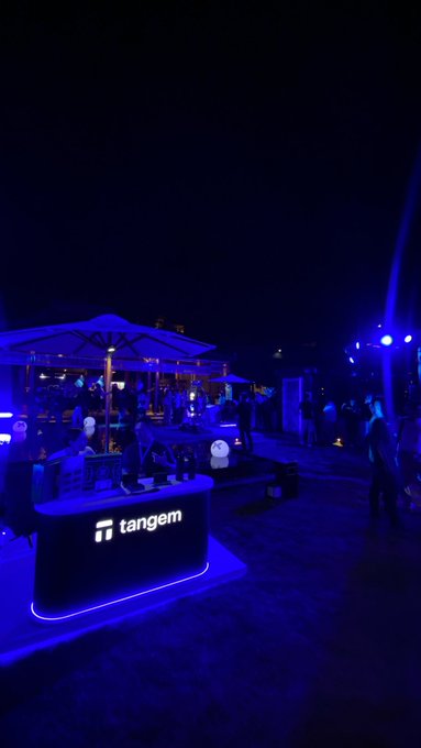 An evening outdoor event features a Tangem-branded bar illuminated with blue lighting under umbrellas.