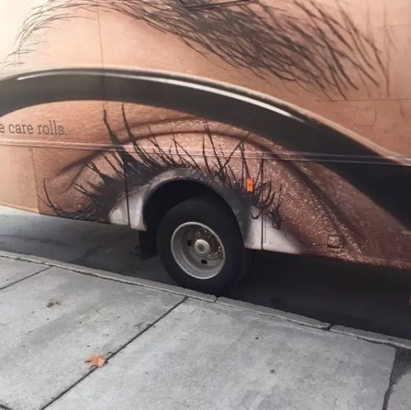 A bus parked on a street featuring a large advertisement showing a close-up of an eye and eyebrow, with partial text reading 'care rolls'.