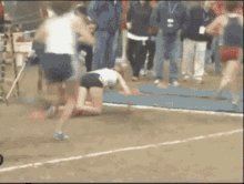 The image shows a person running across a track field during an event or competition. The runner appears to be in motion, possibly falling over as they run. There are several other people present in the scene, some of whom might also be participating in the same event or simply observing it. A few individuals can be seen standing on the sidelines, likely watching and cheering for their fellow competitors.