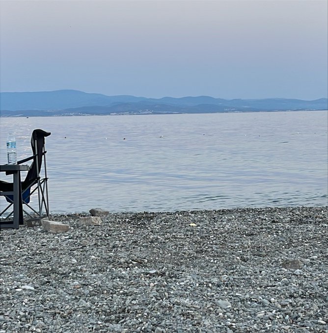 A serene seascape viewed from a rocky pebble beach, featuring a black folding chair and a water bottle in the foreground, with calm waters stretching toward distant blue mountains under a pale sky.