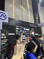 A person's hand making a peace sign in the foreground at what appears to be a trade show or convention, with a crowd of attendees and booths visible in the background.