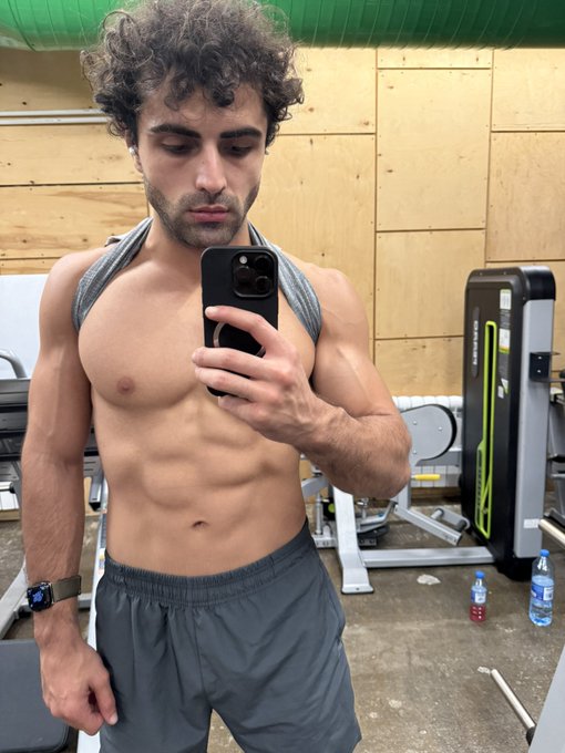 A shirtless, muscular man with curly hair takes a mirror selfie in a gym using a black iPhone, wearing gray shorts and a watch.