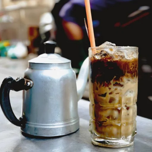 A tall glass of iced coffee with milk layered over ice next to a silver metal kettle on a metallic surface.