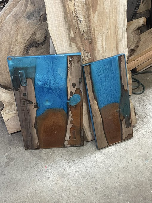 Two large pieces of wood are decorated with vibrant blue resin and brown/amber accents, forming rectangular art pieces displayed against live-edge wood slabs in a workshop setting.