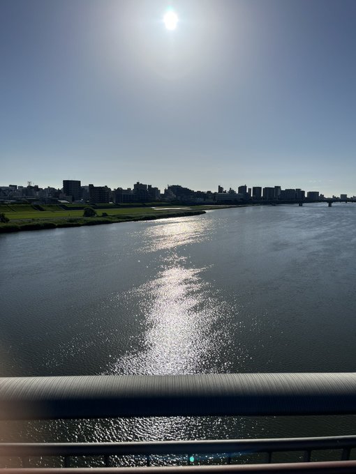 The image shows a beautiful view of a river with a city skyline in the background, including buildings that are reflected in the water's surface. The sun shines brightly over the scene, creating a warm glow on the water and highlighting the architectural details of the cityscape. The photo is taken from an elevated position, providing a unique perspective of both the river and the city below it.