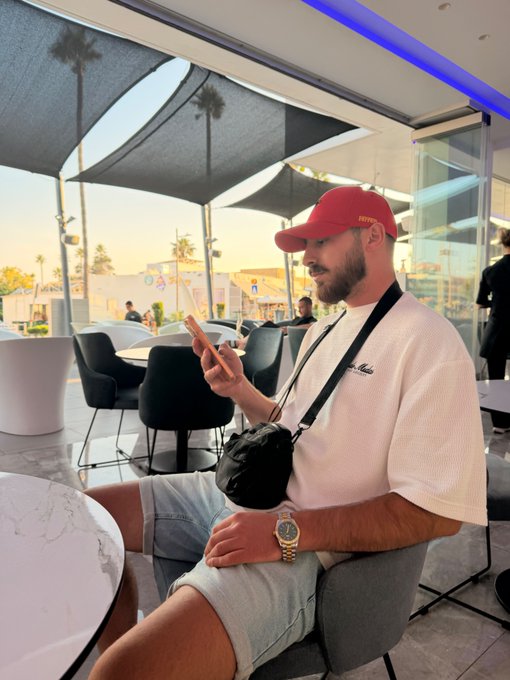 A man wearing a red Ferrari cap and white t-shirt sits at an outdoor café, checking his phone while wearing a gold watch and a black crossbody bag. The casual morning setting and relaxed vibe match the "Gm friends" greeting, capturing a leisurely start to the day.