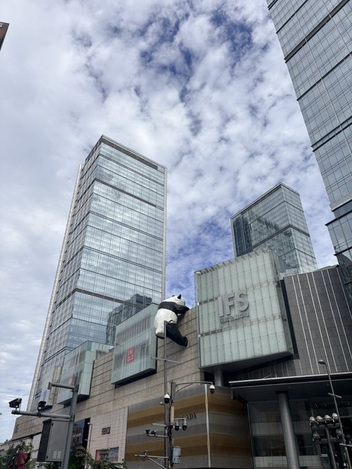 A modern urban scene features tall glass skyscrapers under a cloudy sky, with a large panda sculpture climbing a building labeled 'IFS'.