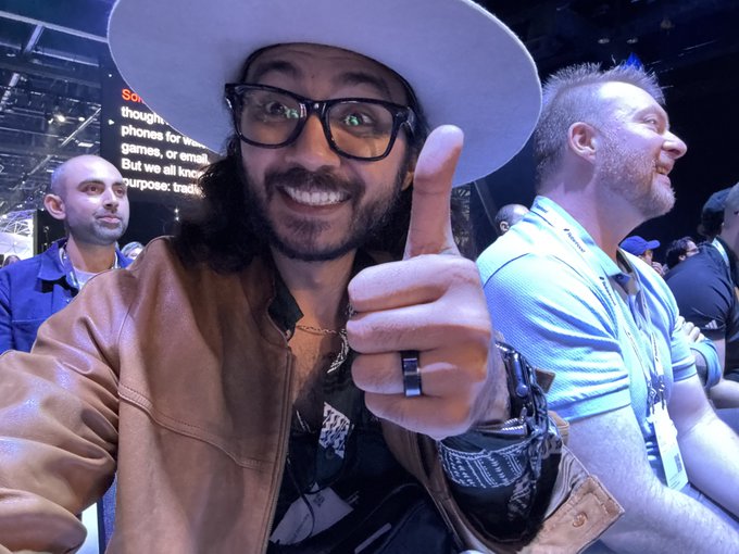 A bearded man wearing black-rimmed glasses, a white wide-brimmed hat, and a brown leather jacket gives a thumbs-up and smiles at the camera at what appears to be a convention, with another attendee in a blue shirt visible beside him.