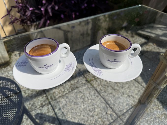 The image features two cups of coffee sitting side by side on a glass table, both placed on saucers. The background includes some plants and a chair, creating an outdoor setting for enjoying a cup of coffee. The caption reads "no better way to start the week than with a coffe in good company waiting for the markets to moon back from the lows". This message suggests that the two cups of coffee are meant to be enjoyed together as a symbol of friendship or camaraderie, and that they should be savored while discussing current events or sharing thoughts on how to navigate the market's fluctuations.