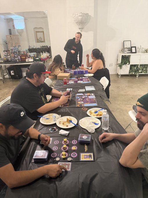 A group of people are gathered around a long table playing a card game, with plates of food, drinks, and a 'Spirit Hunters' game box on the table.