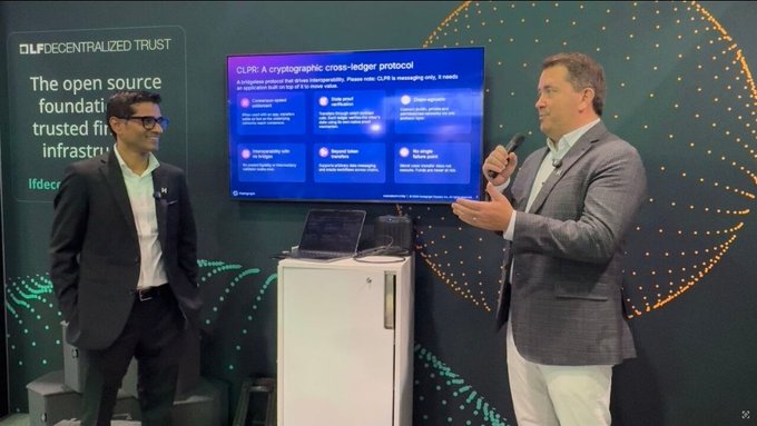 Two men in business attire presenting at a technology conference, with one speaking into a microphone. A screen displays 'CLPR: A cryptographic cross-ledger protocol' and a banner reads 'LF DECENTRALIZED TRUST'.