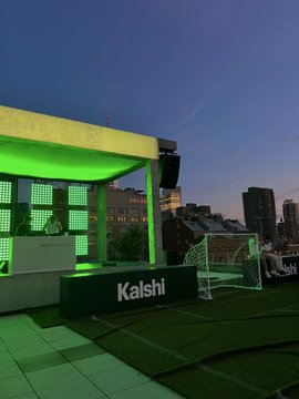 An evening view of a rooftop bar or event space branded with 'Kalshi', featuring green illuminated structures and a city skyline in the background.