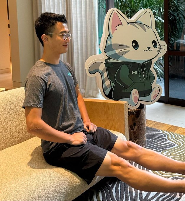 A man wearing glasses and a gray t-shirt sitting on a sofa, smiling and looking at a cutout of a cartoon cat character wearing a green hoodie, in what appears to be a modern home interior.