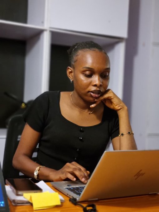 A young woman in a black button-up top works intently on a silver laptop at a desk, accessorized with gold jewelry including a clover bracelet, watch, and necklace.