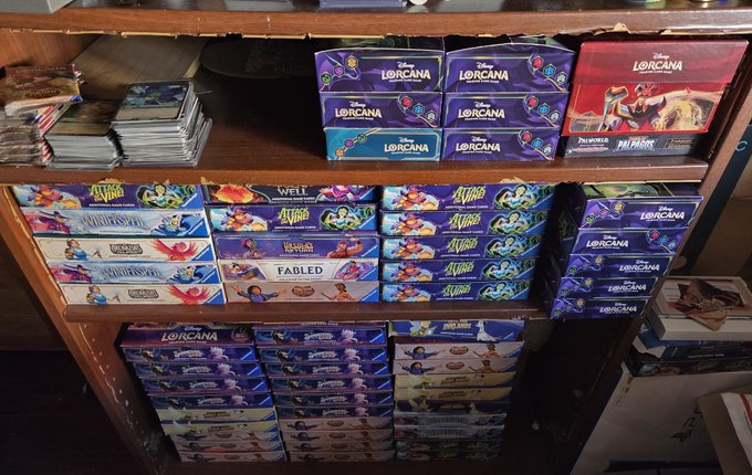 A retail display shelf holds stacked bulk boxes of Disney Lorcana trading card game booster boxes, including sets labeled "Fabled," "Wellspring," and "Promos," alongside loose booster packs, with handwritten signage on the side indicating "Bulk" sales.