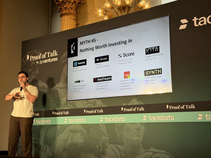 The image features a man standing behind a podium, giving a speech at an event. The podium has a large screen displaying information about investing in companies like IOTA, SynthData Co., and others. The speaker appears to be discussing the Myth of Hacking and its implications for investing in tech companies.