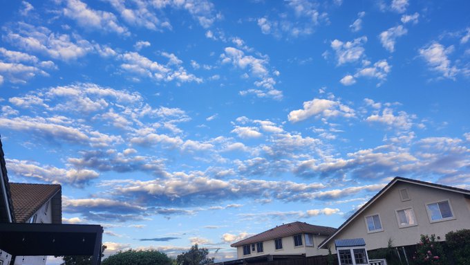 A bright blue sky filled with scattered, fluffy white clouds sits above the rooftops of suburban houses with beige and white walls.