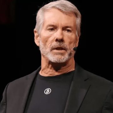 A man with gray hair and a beard, wearing a dark blazer over a black t-shirt with a Bitcoin logo, speaks at what appears to be a presentation.