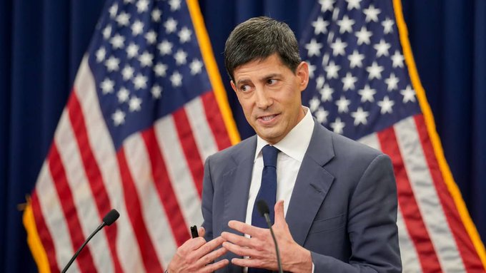 A man in a dark suit and tie speaks at a podium flanked by two American flags, gesturing with his hands as he addresses an audience. The image accompanies a post about Federal Reserve Chair Kevin Warsh raising interest rates by 25 basis points to combat persistent inflation, with the speaker's serious demeanor reflecting the gravity of the hawkish policy decision.