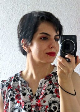 A mirror selfie of a person with short dark hair wearing red lipstick and a floral patterned shirt, holding a small camera.