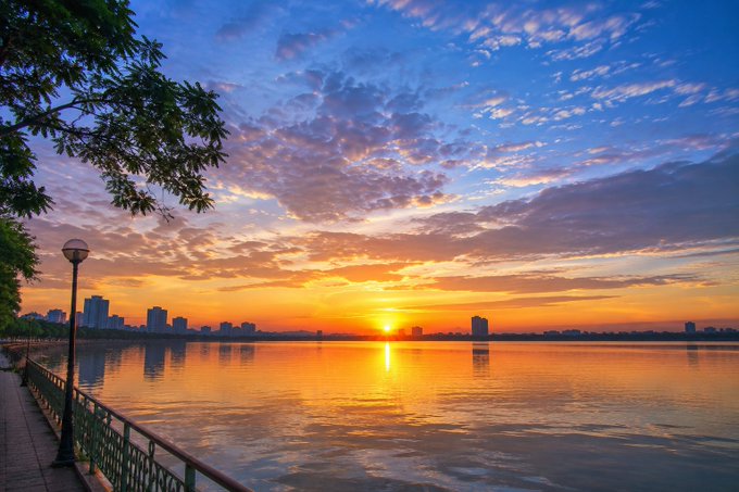 A breathtaking sunrise over a tranquil riverside, with the sun's golden glow reflecting on calm waters beneath a sky painted in warm orange and soft blue hues, while a city skyline silhouettes the distant horizon. The image captures the fresh, hopeful energy of a new week, perfectly complementing the uplifting message of starting the week with positive energy, smooth work progress, and an enthusiastic push to build and trade "X pay" stocks stronger.