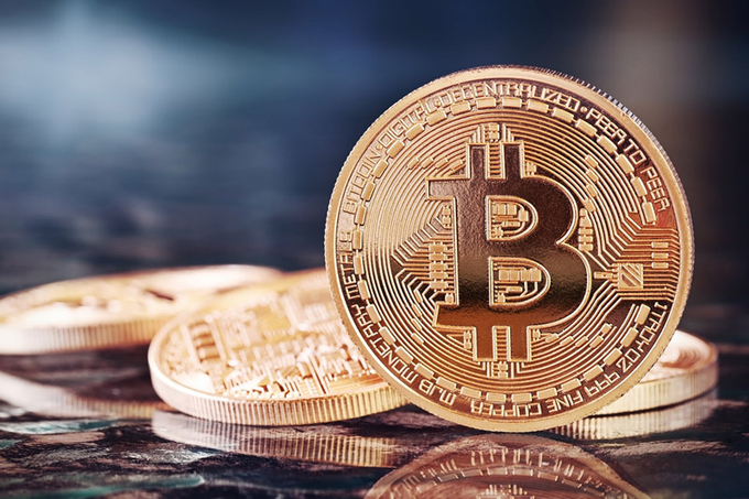 A close-up of a gold-colored Bitcoin coin resting against other similar coins on a dark, reflective surface.