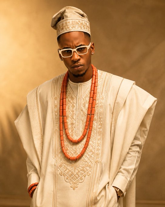 A man in traditional Nigerian attire featuring a white embroidered agbada, coral bead necklaces, white-framed glasses, and a matching cap.
