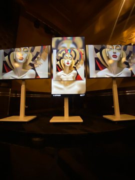 Three framed art pieces displayed on wooden stands in a dimly lit setting, featuring stylized portraits of women with bold geometric designs in red, black, and white color schemes reminiscent of Art Deco or Cubist styles.