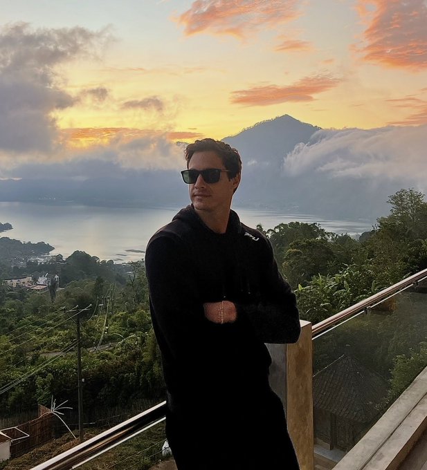 A man wearing sunglasses and a black hoodie stands on a balcony overlooking a scenic lake and mountain landscape at sunset.