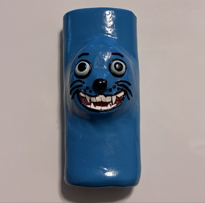 A blue ceramic or clay sculpture featuring a wide, grinning cat-like face with prominent teeth and large eyes, mounted against a plain white background.