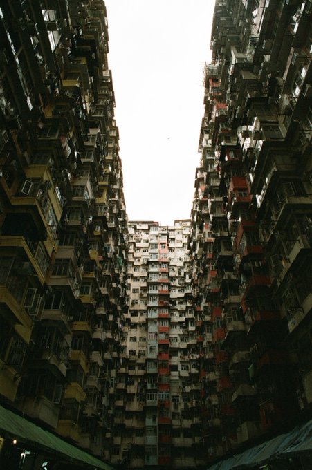 A low-angle photograph captures the dense, towering facade of a residential building complex, likely the Yick Cheong Building in Hong Kong, with countless balconies and air conditioning units reaching toward a sliver of sky.