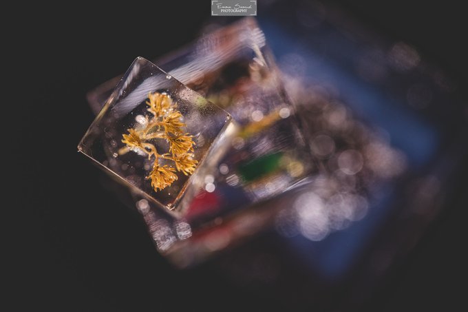 A macro photograph by Eman Saeed Photography showing a clear acrylic cube containing golden crystalline structures, with colorful bokeh lights reflecting on its surface.