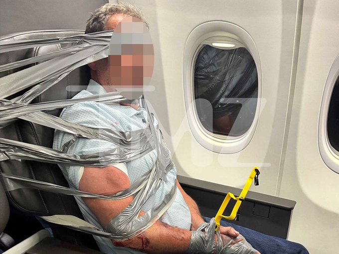 A photo showing a man seated in an airplane seat wrapped extensively in duct tape around his torso and wrists, with duct tape also covering his mouth and eyes, while holding a yellow plastic tool.