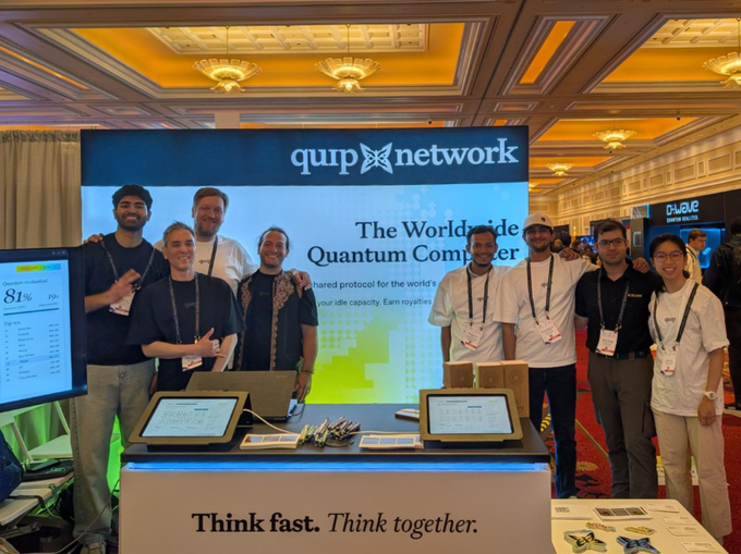 A group photo of the Quip Network team at a conference or trade show booth promoting 'The World's Wide Quantum Computer,' with the tagline 'Think fast. Think together.' displayed on the booth counter.