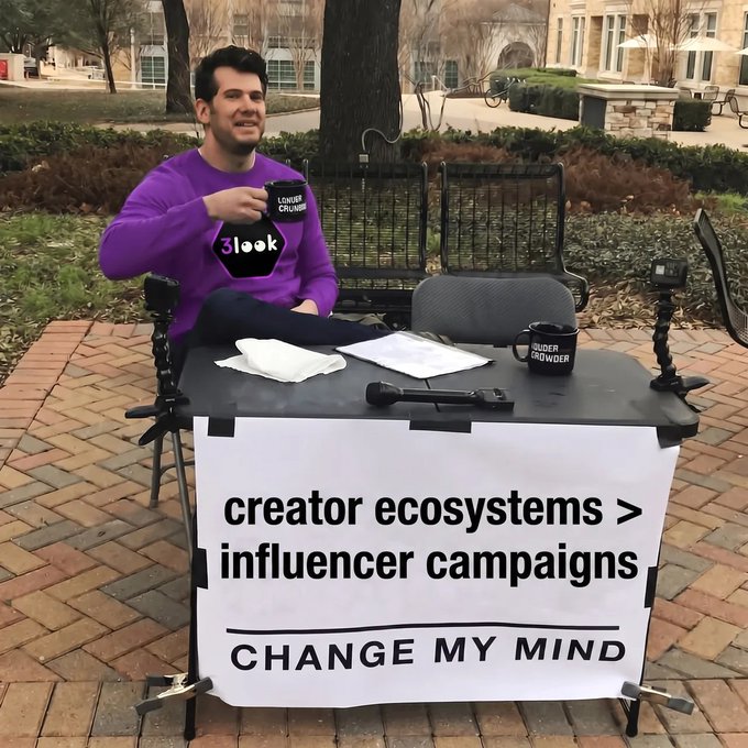 The image shows a man sitting behind a desk with a sign that says "Change My Mind" above him. The sign also has a question about creator ecosystems and influencer campaigns, which seems to be related to the content of the post on social media. The man is holding a cup in his hand, possibly enjoying a beverage while reading or writing something down at the desk.