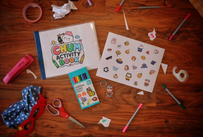 A flat lay of stationery and craft supplies on a wooden table, including a 'CHUM ACTIVITY BOOK', colored pencils, stickers, scissors, and tape.