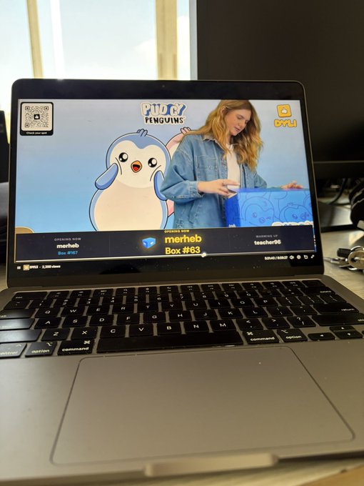A MacBook displays a YouTube video titled "Pudgy Penguins" featuring a blonde woman unboxing a blue "merheb Box #83" mystery box alongside a cute penguin cartoon mascot. The scene captures the "shouting" moment referenced in the post, as the woman excitedly reveals her highly anticipated Pudgy Penguins collectible unboxing.