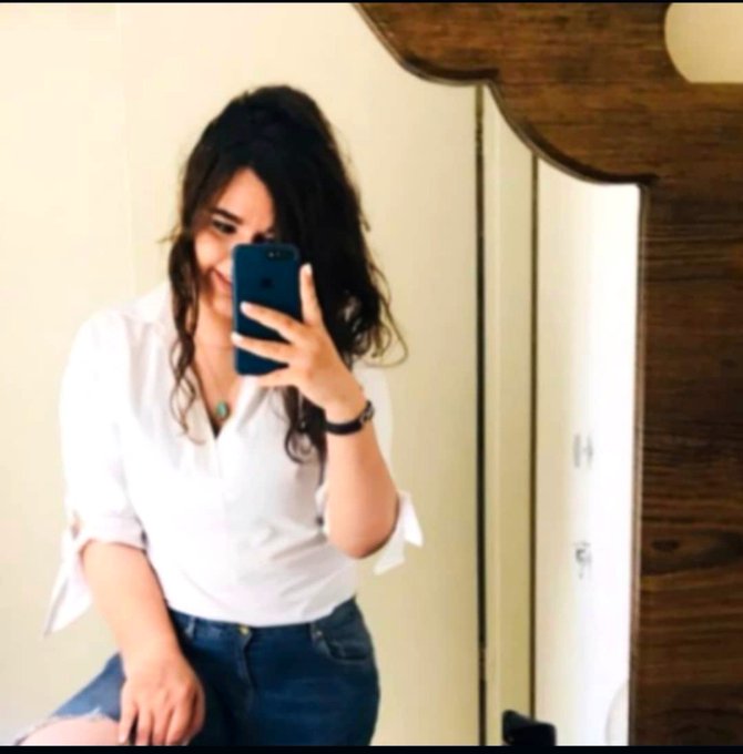 A mirror selfie of a woman with long dark hair wearing a white button-up shirt and denim shorts, taking the photo with a blue smartphone in what appears to be a bedroom with a wooden mirror frame.