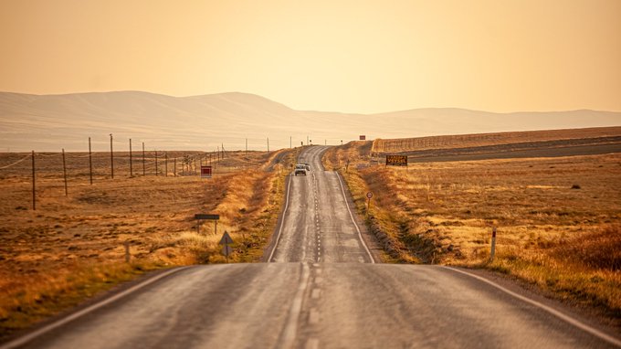 A long, empty road stretches into golden-hued hills at sunset, with a single small car visible in the distance, evoking a sense of endless journey that mirrors the song's title "Until the End."
