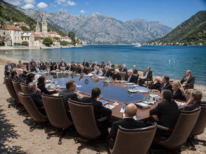 A wide shot of world leaders seated around a circular conference table on a pebble beach, with calm blue water and mountains in the background.