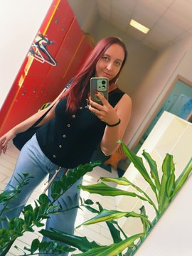 A mirror selfie of a young woman with pink-tinted hair wearing a black vest and jeans, holding a smartphone, with green plants visible in the foreground.
